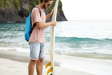 Stylish young Caucasian male surfer with blue bag examining his white surfboard before riding powerful waves, standing on sandy beach with vast ocean and rocky cliff with vegetation behind him