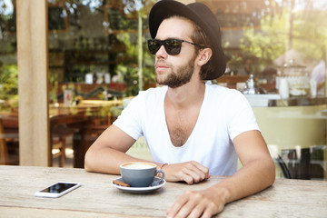 Fototapeta na wymiar People, lifestyle, travel and vacations concept. Handsome bearded tourist wearing stylish hat and sunglasses sitting at wooden table with cell phone and mug during breakfast at sidewalk cafeteria