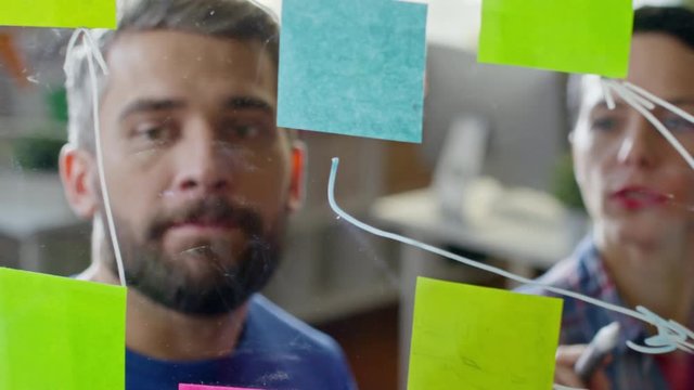 Close up of creative startup workers looking at glass wall planner with sticky notes discussing ideas and drawing arrows. Suitable for business, teamwork, innovation, project planning