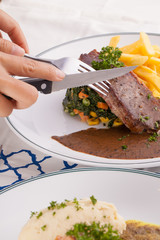 A female hand using knife cutting beef steak decorated with fries in a nice dish set on a table.