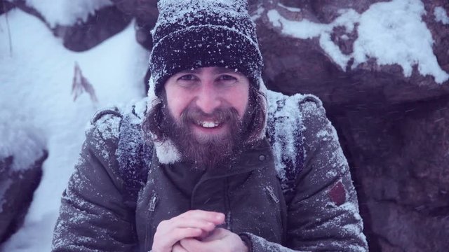 Young bearded man in snowbound clothes warming up his hands after snowfall on rocky outdoors background