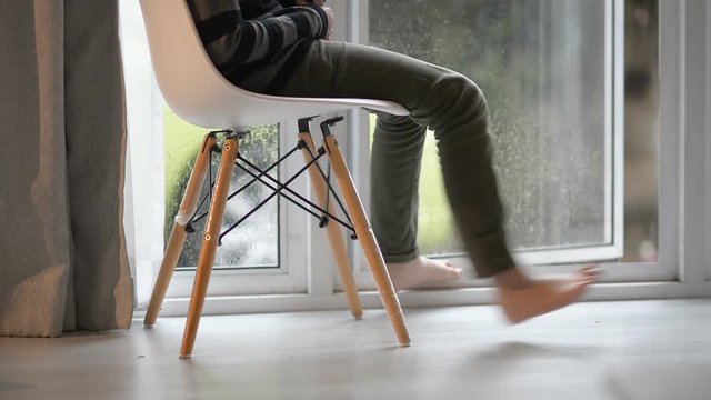 Boy shaking legs while use tablet and sitting near window at home in rainy day