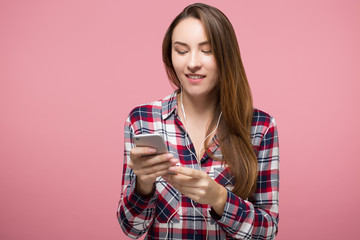 hipster girl listening to the music in in-ear headphones and going through the playlist in her phone over pink background isolated