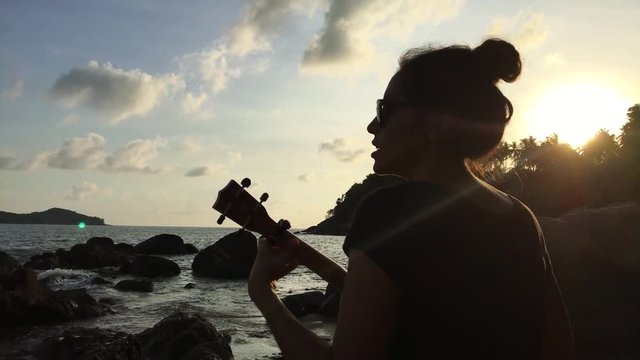 Young mixed race girl playing the ukulele on the beach rocks. Colorful sunset. HD back view silhuette. Thailand.