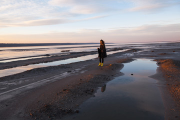Beautiful girl is standing on the sea in a Sunbeam, yellow boots, long black coat, sand underfoot