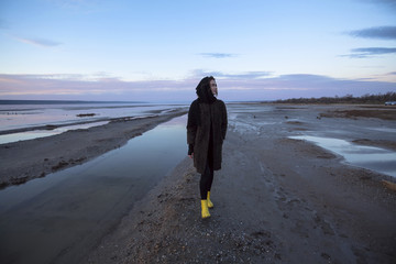 Beautiful girl is standing on the sea in a Sunbeam, yellow boots, long black coat, sand underfoot
