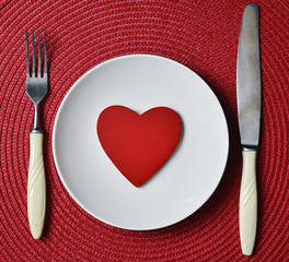 St.Valentine's Day romantic dinner concept with white plate, knife, fork and decorative wooden heart on red straw napkin background.Selective focus.