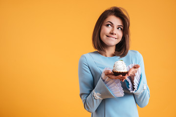 Pensive smiling young woman holding cupcake and thinnking - obrazy, fototapety, plakaty