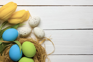 Easter background with colorful eggs in nest and yellow tulips over white wood. Top view with copy space