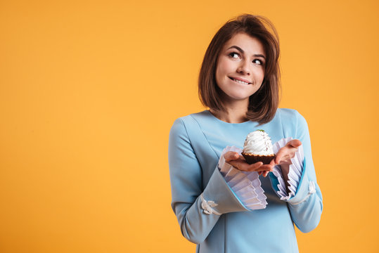 Pensive smiling young woman holding cupcake and thinnking