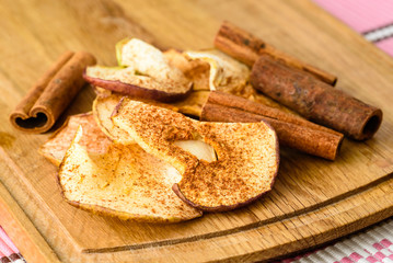 Dehydrated apple slices with powdered cinnamon and whole cinnamon on wooden cutting board.