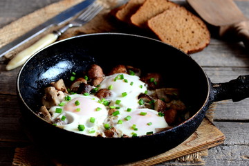 Fried eggs with mushrooms in pan. Brown bread slices, fork, knife, cutting board on a wooden table. Vegetarian easy egg breakfast. Homemade recipe. Vintage style. Closeup