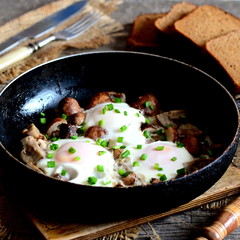 Fried eggs in pan. Fresh eggs fried with mushrooms and green onions. Brown bread slices, fork, knife, cutting board on a wooden table. Healthy egg breakfast. Homemade recipe. Rustic style. Closeup