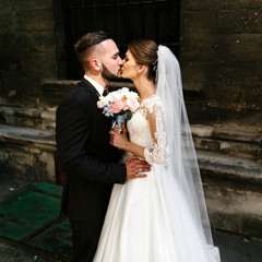 Tender kiss of dreamy wedding couple posing before wall of old c