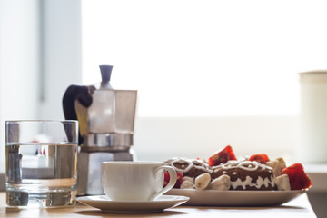Holiday breakfast in home kitchen. Espresso cup, cookies and sweets, glass of water and italian moka coffee pot on natural wood table served for a christmas breakfast in front of window with sunlight