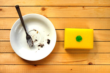 Dirty white plate in the white sink in the kitchen and yellow dish sponge with a drop of dish-washing detergent