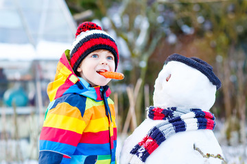 Funny kid boy in colorful clothes making a snowman, outdoors