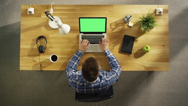 Top View of a Young Creative Man Working on His Green Screen Mock-up Laptop while Sitting at His Wooden Desk. Also on the Table: Coffee Cup, Smartphone, Notebook, Lamp, Plant.  