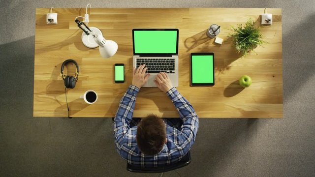  Top View of a Creative Young Man Sitting at His Desk Typing on Laptop Computer Beside Lies Smartphone and Tablet Computer, All Electronic Devices Have Green Mock-up Screen. 