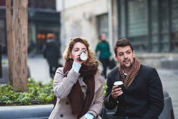 Young couple with coffee to go in hands siiting on bench in urban street. 