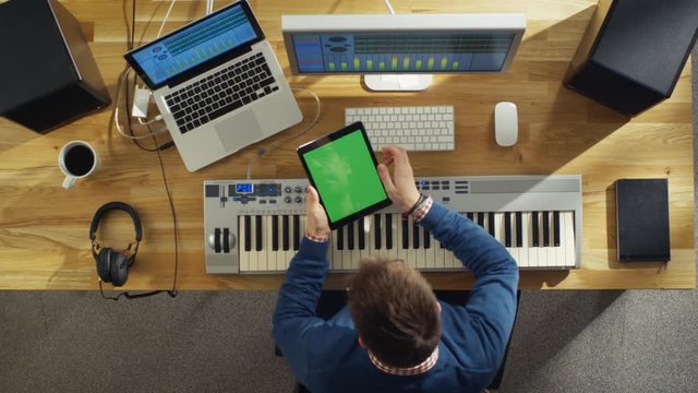 Top View of a Musician Holding Green Screened Tablet Computer While Working in His Studio. His Workspace is Hi-Tech but also Sunny and Warm Looking.