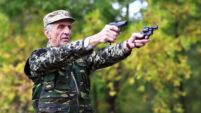 Soldier in military uniform shoots from two pneumatic handguns. Senior man shoots a two pistols in forest. Soldier in military uniform shoots a revolver. Retired officer at shooting range