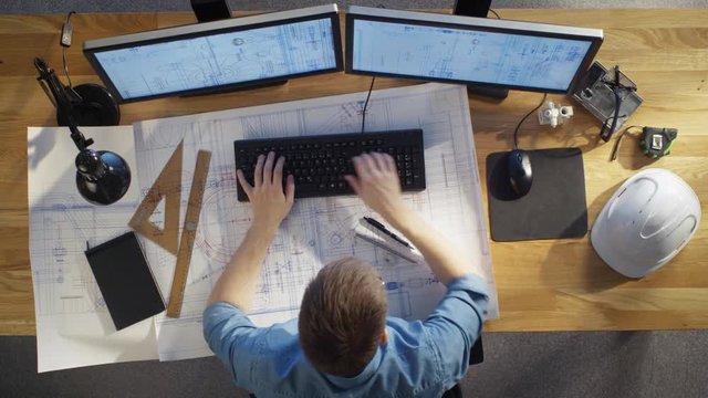  Top View of architectural Engineer Working on His Blueprints, Holding Tablet Computer, Using Desktop Computer Also. His Desk is Full of Useful Objects and Evening Sun.
