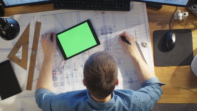 Top View of Architectural Engineer Draws on His Blueprints, Compares with Tablet Computer with Green Screen, Using Desktop Computer Also. His Desk is Full of Useful Objects and Morning Sun. 