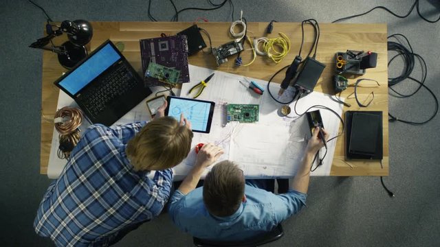  Top View of a Professional IT Technician Soldering Circuit Board. He's Interrupted by Colleague Holding Tablet Computer. They Both Discuss Blueprint Shown on Tablet. 