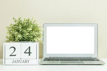 White wooden calendar with black 24 january word with white blank space at center of computer notebook on white wood desk and cream wallpaper textured background , selective focus at the calendar