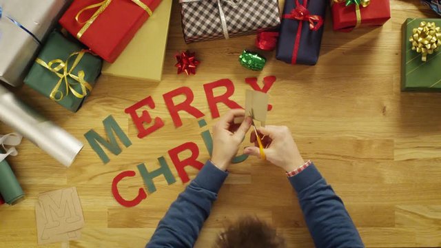 Top View Time-Lapse of a Man Cutting Letters for Words "Merry Christmas" and laying Them on The Wooden Table With Gift Wrapped Boxes.  Shot on RED Cinema Camera in 4K (UHD).
