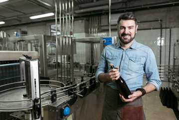 Smiling man holding a bottle of alcohol at brewing factory