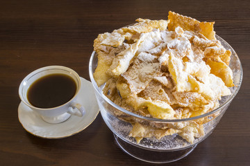 Sprinkle with powdered sugar, fried plate of shortcake dough. Polish cuisine - faworki ( angel wings) .  Faworki in a glass vase and coffee in a porcelain cup on a dark background..