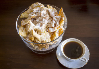 Sprinkle with powdered sugar, fried plate of shortcake dough. Polish cuisine - faworki ( angel wings) .  Faworki in a glass vase and  coffee in a porcelain cup on a dark background.