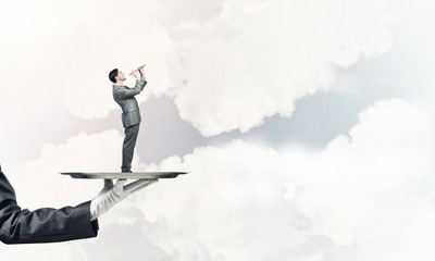 Businessman on metal tray playing fife against blue sky background