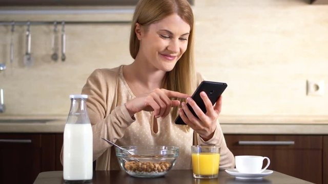 Beautiful young attractive woman having cornflakes for breakfast and using her smartphone.