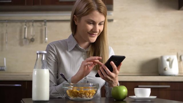 Beautiful young attractive businesswoman having cornflakes for breakfast and using her smartphone.