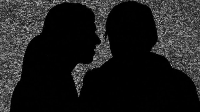 A woman whispering a secret in the ear of a man. Silhouette shot over the fuzzy noise of an old tv screen.
