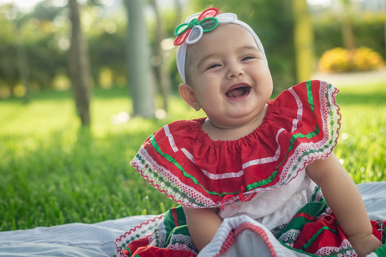 White baby dressed in the colors of the Mexican flag, sitting on green grass, spring, with emotional expression, happy, original. Red, green and white dress. White ribbon on the head and three-colored