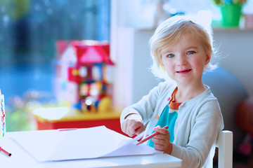 Cute little left-handed child is drawing and painting with colorful felt-tip pens. Preschooler girl creating at home or kindergarten sitting at small table in bright sunny playroom