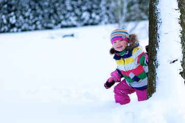 Portrait of beautiful toddler girl playing outdoors with snow. Happy little child wearing colorful knitted hat and blue coat enjoying winter day in the park or forest.