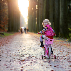 Little child riding her bicycle in the park. Cute preschooler girl learning to cycle with stabilisers wheels. Sportive kid enjoying sunny day outdoors in the forest.