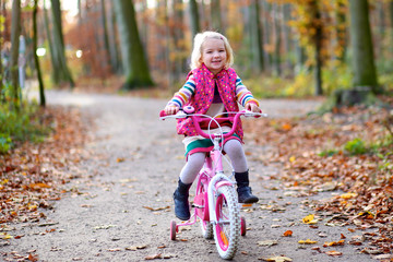 Little child riding her bicycle in the park. Cute preschooler girl learning to cycle with stabilisers wheels. Sportive kid enjoying sunny day outdoors in the forest.
