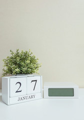 White wooden calendar with black 27 january word with clock and plant on white wood desk and cream wallpaper textured background , selective focus at the calendar