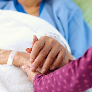 Caring hands holding kind elderly lady's hands in bed at hospital