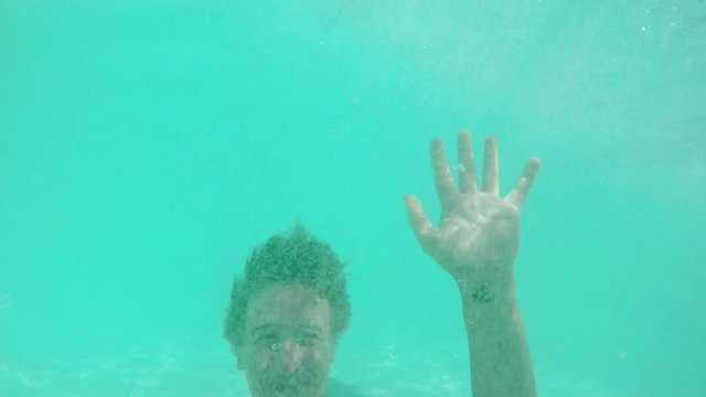 Topless Caucasian male with light beard swimming laughing smiling showing ok in pool. Underwater footage in slowmotion