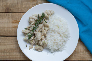 White plate with rice and sliced chicken breasts cooked with cream and decorated with fresh rosemary. Chicken stroganoff served on wooden table with blue napkin