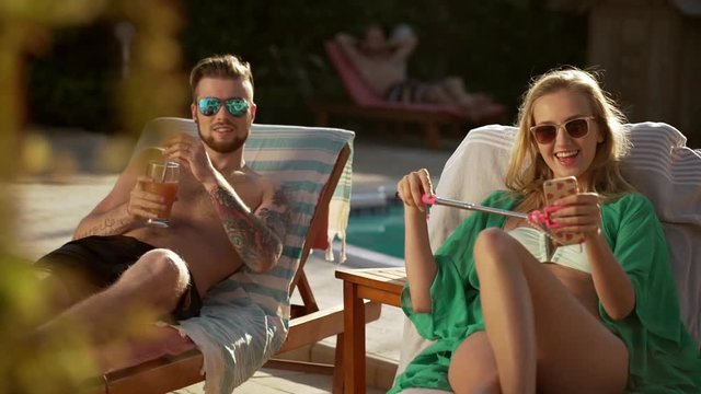 Beautiful Caucasian pair lying in lounges in sunglasses drinking cocktails with mobile phone and selfie stick near pool. In slowmotion