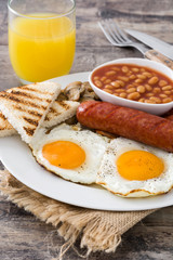 Traditional full English breakfast with fried eggs, sausages, beans, mushrooms, grilled tomatoes and bacon on wooden background
