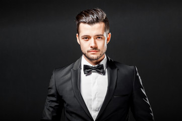portrait of confident handsome man in black suit with bowtie posing in dark studio background